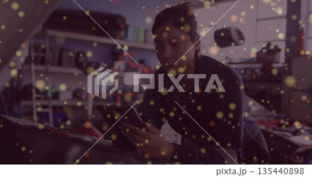 Leaning woman mechanic consulting smartphone under car hood at repair shop, with tool shelves Leaning woman mechanic consulting smartphone under car hood at repair shop, with tool shelves 135440898