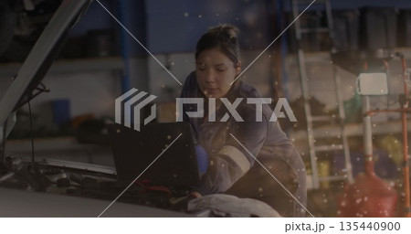 Leaning female mechanic in blue coverall diagnosing engine at repair garage, with laptop on engine 135440900