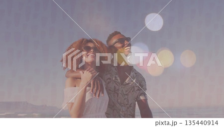 Smiling couple wearing off-shoulder dress, floral-shirt and sunglasses standing on beach by ocean 135440914