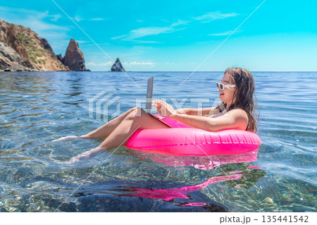 Freelancer laptop beach woman works on computer while floating in pink ring on clear ocean water during summer representing digital nomad lifestyle 135441542