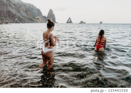 Women, ocean, beach. Two women wade into clear water at a rocky beach with distant mountains. Women, ocean, beach. Two women wade into clear water at a rocky beach with distant mountains. 135441550