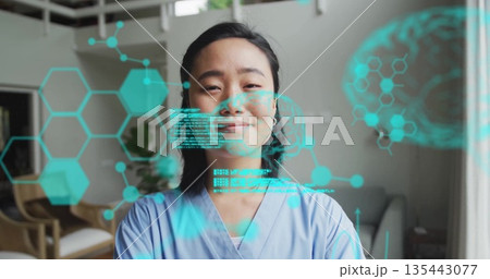 Smiling Korean nurse wearing blue scrubs standing in living room, with holographic medical display Smiling Korean nurse wearing blue scrubs standing in living room, with holographic medical display 135443077