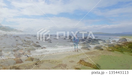 Standing senior hiker wearing jacket and hiking boots on rocky shore, with backpack, poles Standing senior hiker wearing jacket and hiking boots on rocky shore, with backpack, poles 135443113
