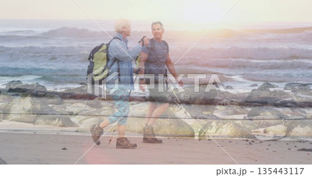 Using trekking poles couple hikers walking along beach at sunrise, with backpacks 135443117