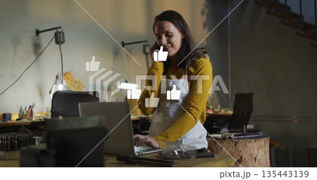 Woman in apron juggling smartphone and typing on laptop in workshop, with thumbs up icons 135443139