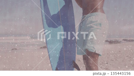 Standing surfer in swim shorts holding light blue surfboard on sandy beach with rocks, copy space Standing surfer in swim shorts holding light blue surfboard on sandy beach with rocks, copy space 135443156