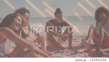 Laughing friends wearing swimwear sitting on beach sand near water's edge, with clear bottled drink Laughing friends wearing swimwear sitting on beach sand near water's edge, with clear bottled drink 135443158