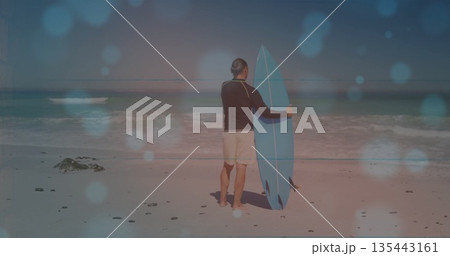 Mature man wearing black rash guard standing on sandy beach with blue surfboard and pebbles Mature man wearing black rash guard standing on sandy beach with blue surfboard and pebbles 135443161