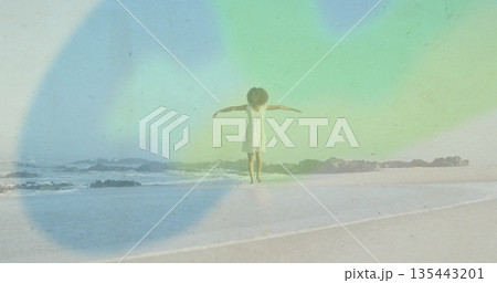 Balancing woman in white sundress spreading arms on beach by waves over rocks, with pastel light 135443201