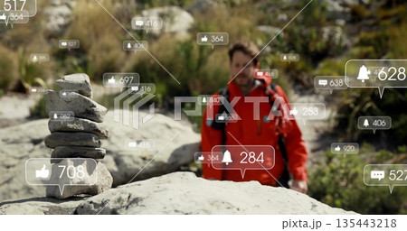 Standing male hiker wearing red jacket on rocky riverbed, with stone cairn and notification icons 135443218
