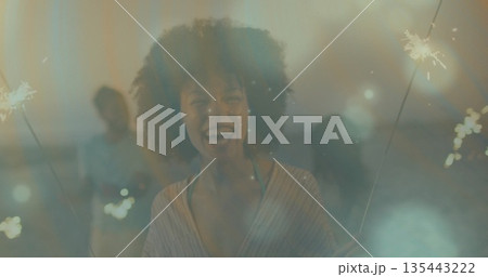 Smiling Black woman wearing striped top holding sparklers on rooftop terrace at night, with friends Smiling Black woman wearing striped top holding sparklers on rooftop terrace at night, with friends 135443222