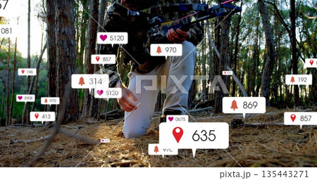 Kneeling man wearing camouflage coat holding compound bow on pine forest floor, with digital alerts 135443271