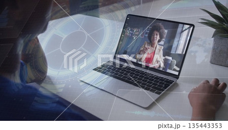 Hosting colleague in tan blazer appearing on laptop screen at home desk, with coffee and notebook 135443353