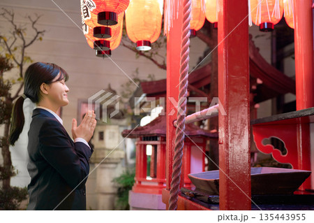 夜の神社で真剣に願い事をするスーツ姿の若い女性 135443955