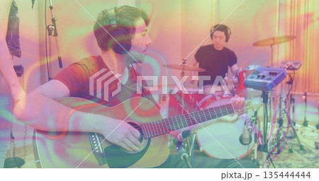 Guitarist strumming acoustic guitar with headphones in studio, drummer playing drum kit, copy space Guitarist strumming acoustic guitar with headphones in studio, drummer playing drum kit, copy space 135444444