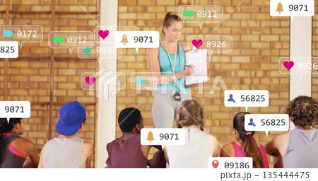 Holding clipboard stopwatch, coach in leggings tank top guiding trainees in gym, social media icons 135444475