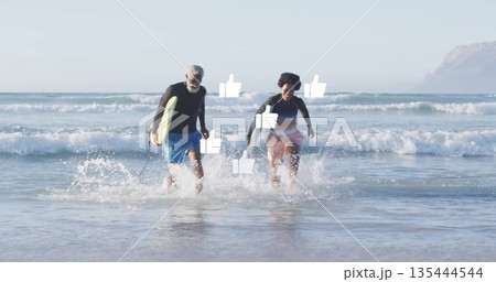 Running senior man and woman carrying yellow surfboard from surf at shoreline, wearing rash guards 135444544