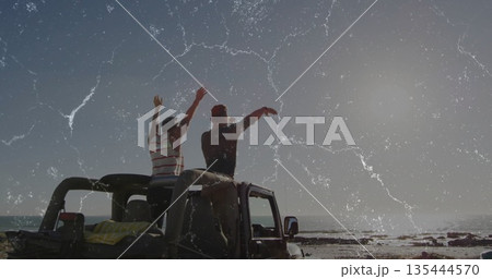 Raising arms, two mature men standing on SUV roof at rocky beach, with towel, ocean Raising arms, two mature men standing on SUV roof at rocky beach, with towel, ocean 135444570