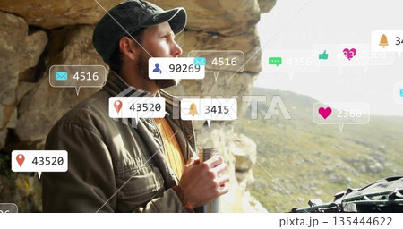 Gazing hiker wearing cap, field jacket at rocky overlook, holding flask and digital overlay icons 135444622