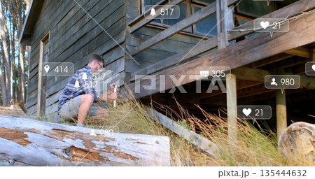 Kneeling male in plaid shirt hammering nails into plank at cabin exterior, with notification icons 135444632