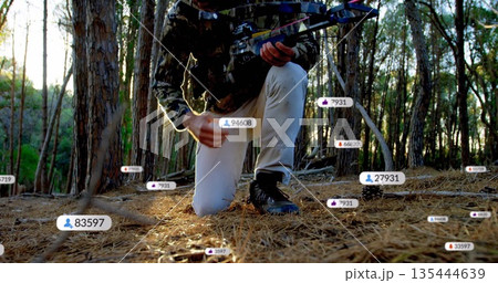 Kneeling hunter in camo clothing handling crossbow in pine forest at sunset, with floating metrics 135444639