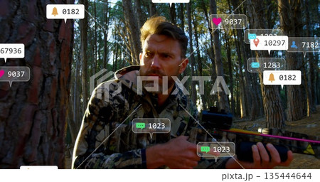 Crouching hunter wearing camo jacket holding crossbow in pine forest, with floating notifications 135444644
