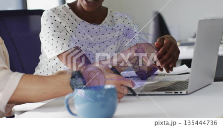 Collaborating businesswomen in suits gesturing toward laptop, taking notes at office with blue mug 135444736