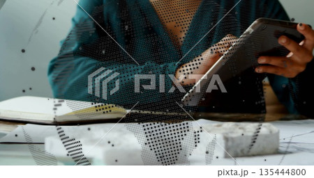 Holding tablet, woman wearing deep teal blouse reviewing design at desk, with model blocks notebook Holding tablet, woman wearing deep teal blouse reviewing design at desk, with model blocks notebook 135444800