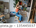 Smiling delighted woman artist stretching arms and body while working on painting at home gallery. 135445037