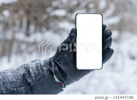 Hand in a black leather glove holding a smartphone with a blank white screen outdoors in a snowy 135445196