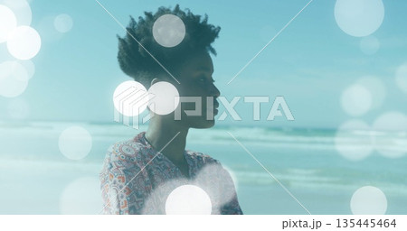 Standing woman wearing paisley blouse gazing toward ocean at beach, with bokeh lights, copy space Standing woman wearing paisley blouse gazing toward ocean at beach, with bokeh lights, copy space 135445464