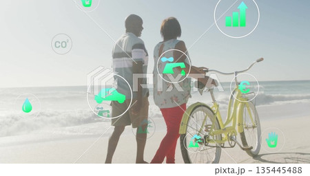 Strolling mature couple pushing yellow bicycle along sandy shoreline, with floating green eco icons Strolling mature couple pushing yellow bicycle along sandy shoreline, with floating green eco icons 135445488