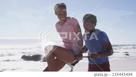 Couple riding and pushing bicycle along beach edge in athletic wear, with world map overlay Couple riding and pushing bicycle along beach edge in athletic wear, with world map overlay 135445494