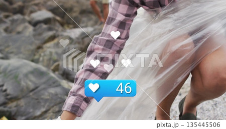 Man wearing flannel wrapping film around her on pebbly beach, with white hearts, blue badge 46 135445506