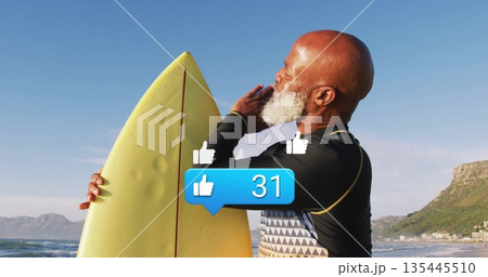 Holding yellow surfboard, senior man in wetsuit top gesturing at beach with cliffside buildings 135445510