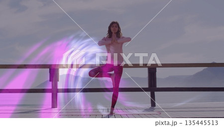Balancing yoga practitioner performing tree pose on wooden pier by water, with digital light swirl 135445513