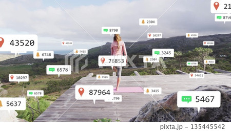 Woman in sportswear performing tree pose on pink yoga mat on valley-view deck, with digital icons 135445542