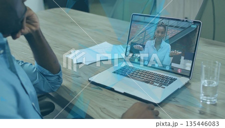 Man wearing light blue shirt leaning on office table, watching call on laptop with documents water Man wearing light blue shirt leaning on office table, watching call on laptop with documents water 135446083