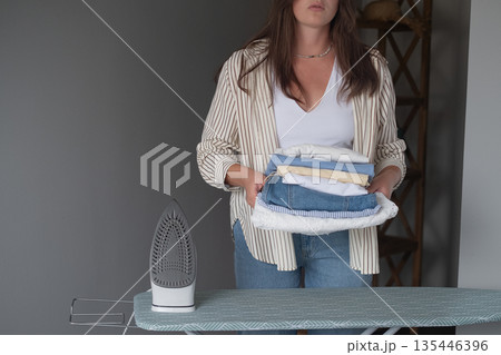Woman irons laundry at home holding a hot iron and neatly stacking clothes showing everyday household duties concept Woman irons laundry at home holding a hot iron and neatly stacking clothes showing everyday household duties concept 135446396