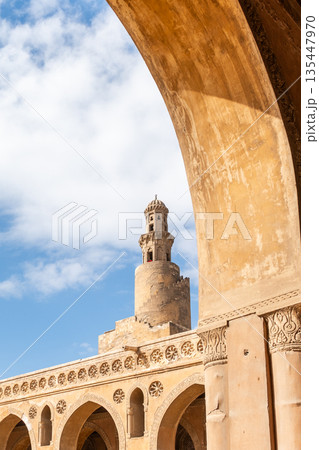 Ibn Tulun Mosque in Cairo viewed through arches. The mosque features a tall minaret and intricate architectural details. Blue sky visible in the background. 135447970
