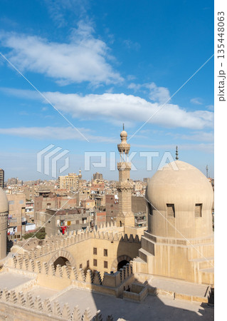View of the Madrasa of Sarghatmish in Cairo. The scene includes domes, a minaret, and surrounding buildings under a blue sky with clouds. View of the Madrasa of Sarghatmish in Cairo. The scene includes domes, a minaret, and surrounding buildings under a blue sky with clouds. 135448063
