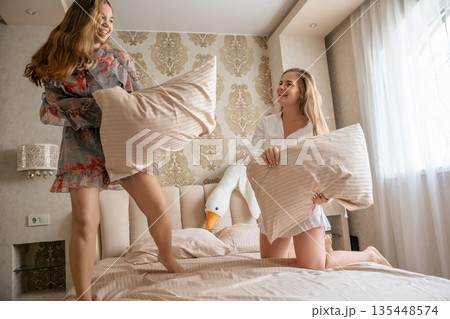 Pillow Fight Bedroom Fun: Two women playfully fight pillows on bed enjoying lighthearted fun in bedroom. Pillow Fight Bedroom Fun: Two women playfully fight pillows on bed enjoying lighthearted fun in bedroom. 135448574