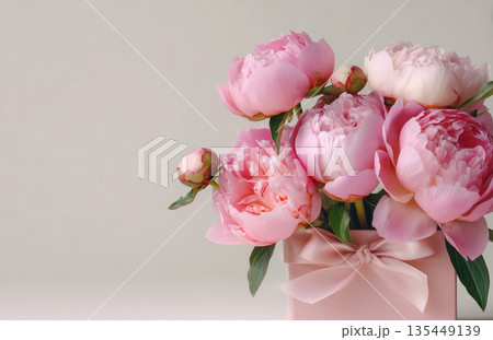 Pink peonies in a square vase with ribbon on plain background in a well-lit room during daytime Pink peonies in a square vase with ribbon on plain background in a well-lit room during daytime 135449139