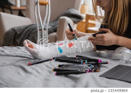 Young teenage girl with broken leg sitting on bed draws with felt tip pens on orthopedic cast. Bored girl with broken leg playing at home during rehabilitation. Accident injury fracture treatment 135449964