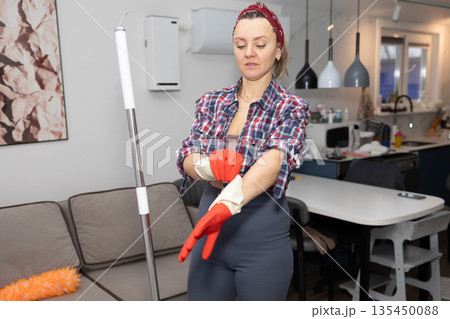 Woman Prepares to Clean Living Room, Putting on Rubber Gloves Woman Prepares to Clean Living Room, Putting on Rubber Gloves 135450088