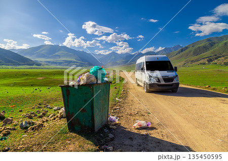 Green trash bin overflowing with garbage on rural dirt road 135450595