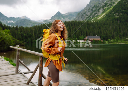 Travel Slovakia, Europe. Tourist with a yellow backpack stands against the backdrop of alpine lake. Travel Slovakia, Europe. Tourist with a yellow backpack stands against the backdrop of alpine lake. 135451493