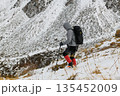 Hiker walking uphill in snowy mountain landscape with backpack and trekking poles. Winter trekking in alpine terrain, cold weather adventure, endurance, and outdoor exploration concept. 135452009