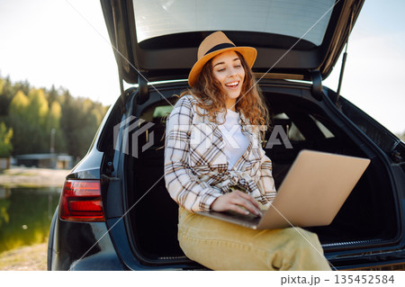 Woman sits on trunk of car, using her laptop while enjoying tranquility of a lake. Remote work. Woman sits on trunk of car, using her laptop while enjoying tranquility of a lake. Remote work. 135452584