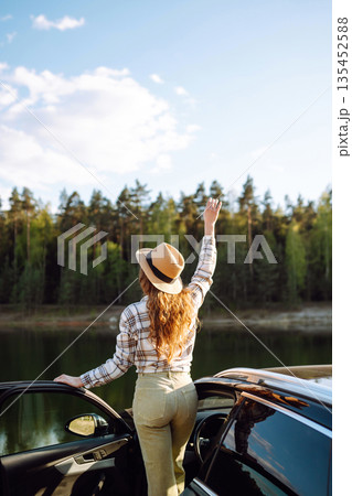 Joyful traveler embraces nature at sunset by lakeside with open car door, freedom, adventure. Joyful traveler embraces nature at sunset by lakeside with open car door, freedom, adventure. 135452588